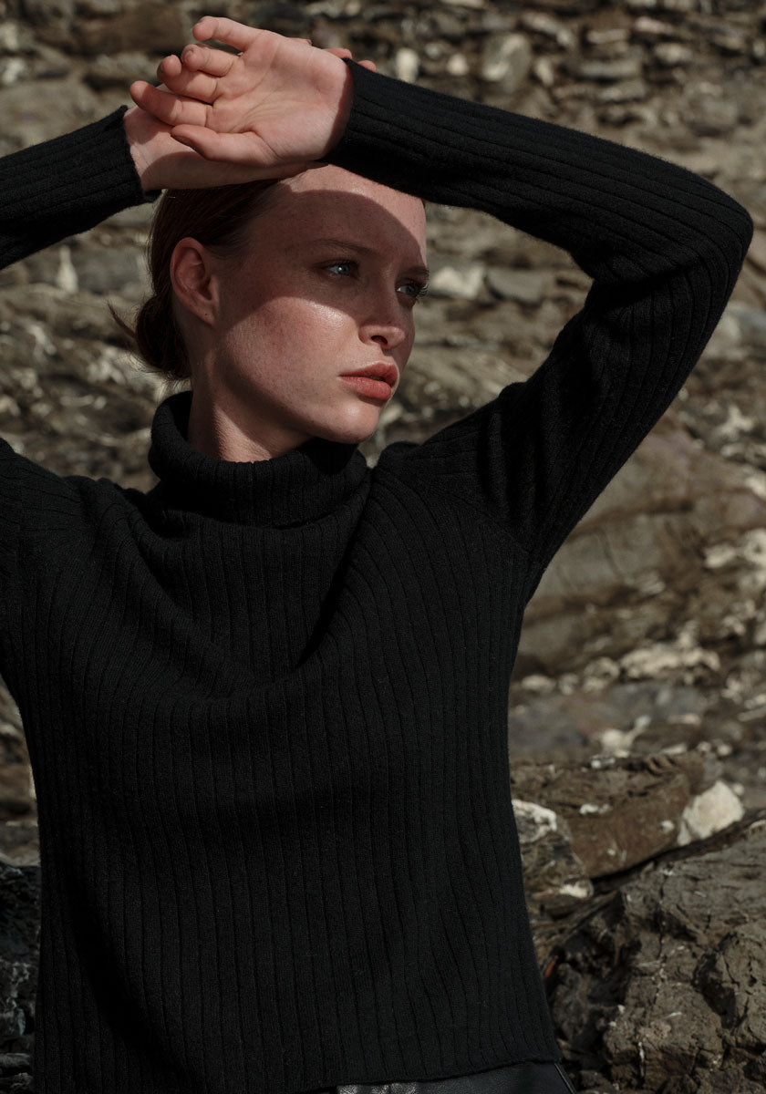 A person in a black Princess Goes Hollywood Rollkragenpullover with ribbed design stands outdoors before rocky terrain, raising both arms to shield their face from sunlight. Their expression is neutral and contemplative, with light casting shadows.