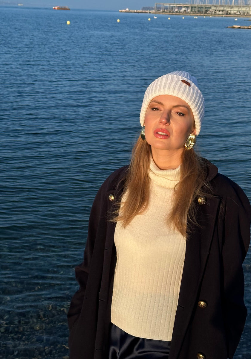 A woman stands by calm water under clear skies, wearing a white knit beanie, large round earrings, and a cream Princess Goes Hollywood Kaschmir Rollkragenpullover mit geripptem Design; sunlight highlights her face.