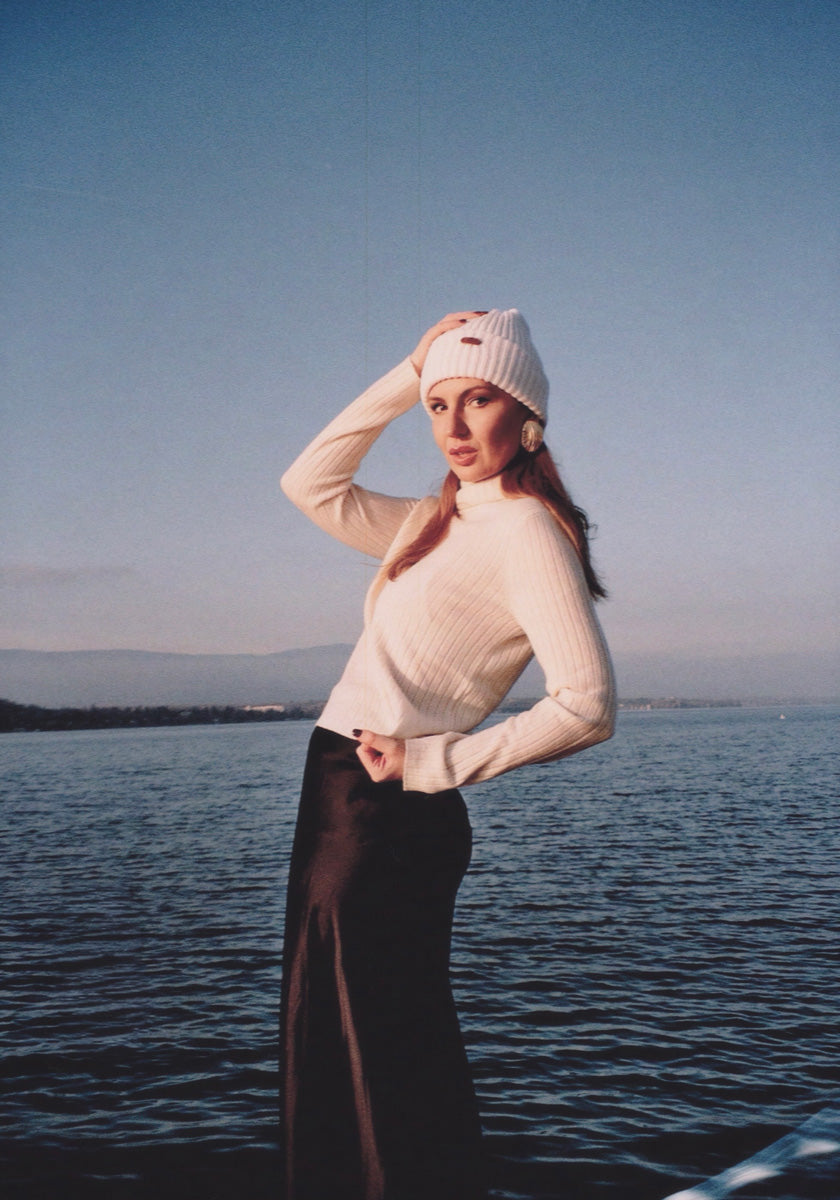 A woman poses by the water wearing a white knit beanie, long black skirt, and a cream ribbed turtleneck sweater from Princess Goes Hollywood. The sky is clear, and distant mountains are visible on the horizon behind her.