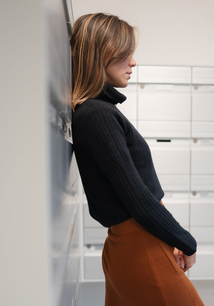 A woman with straight, light brown hair stands in profile by white mailboxes wearing a schwarzen Kaschmir Rollkragenpullover mit geripptem Design von Princess Goes Hollywood und einen rostfarbenen Rock. The minimalist setting enhances the modern vibe.