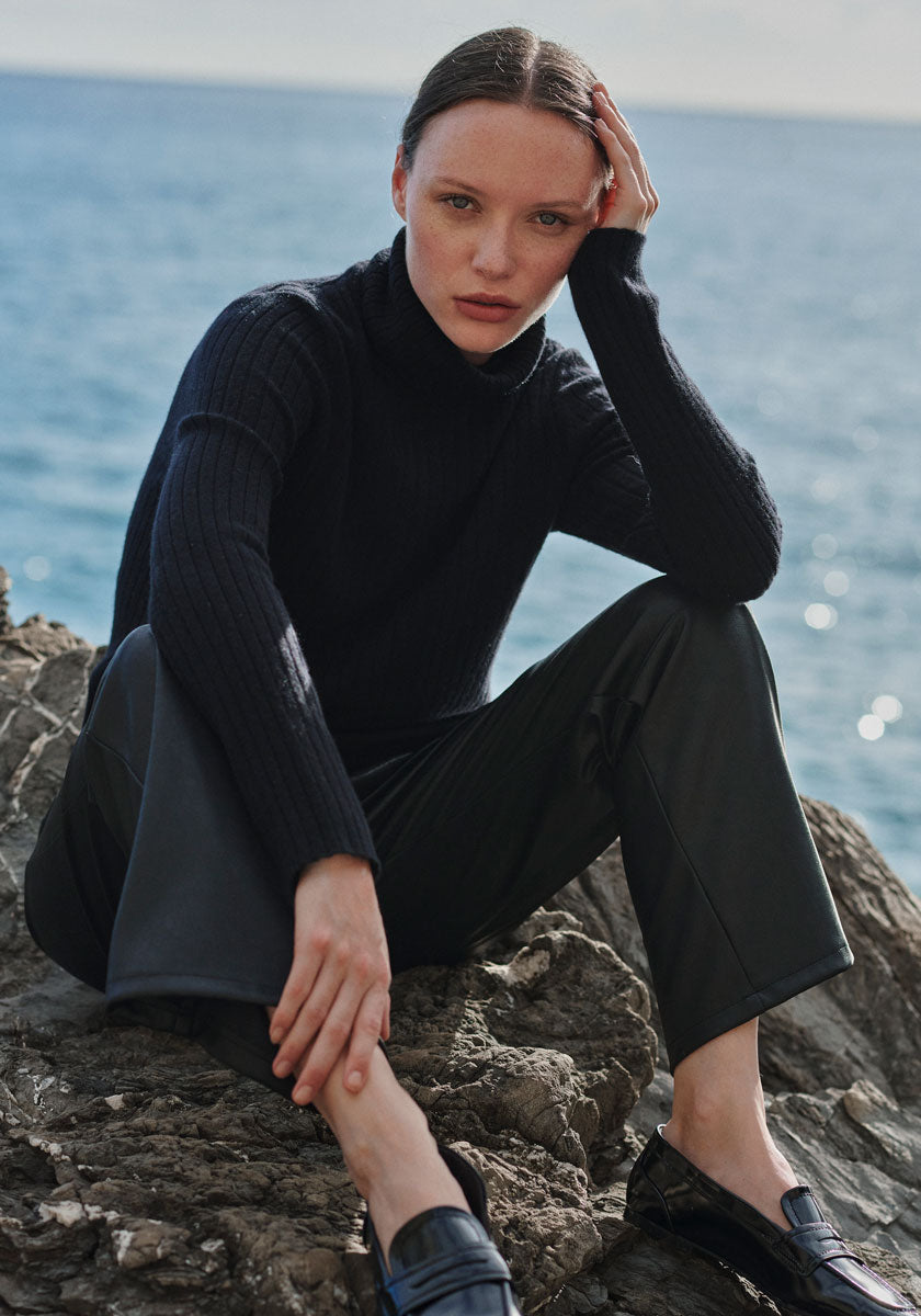 A woman with fair skin and slicked-back hair wears a schwarzen Kaschmir-Rollkragenpullover mit geripptem Design von Princess Goes Hollywood und schwarze Hosen, sitzt auf Felsen am Meer und blickt intensiv in die Kamera, während Wellen im Hintergrund brechen.