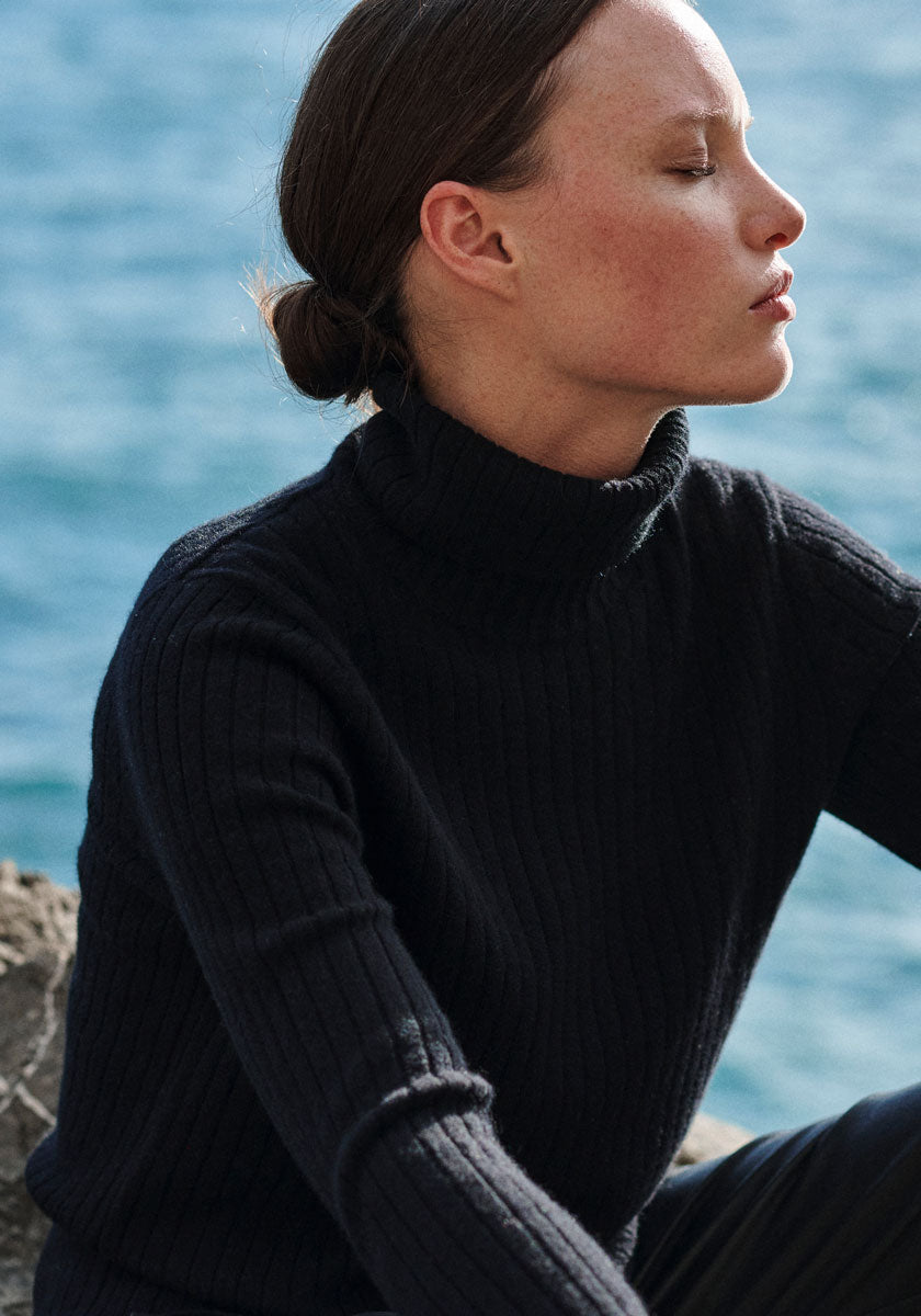 A fair-skinned person with brown hair in a low bun sits outdoors by blue water, wearing a Princess Goes Hollywood geripptes Design Rollkragenpullover. Their eyes are closed and their expression is calm and peaceful.