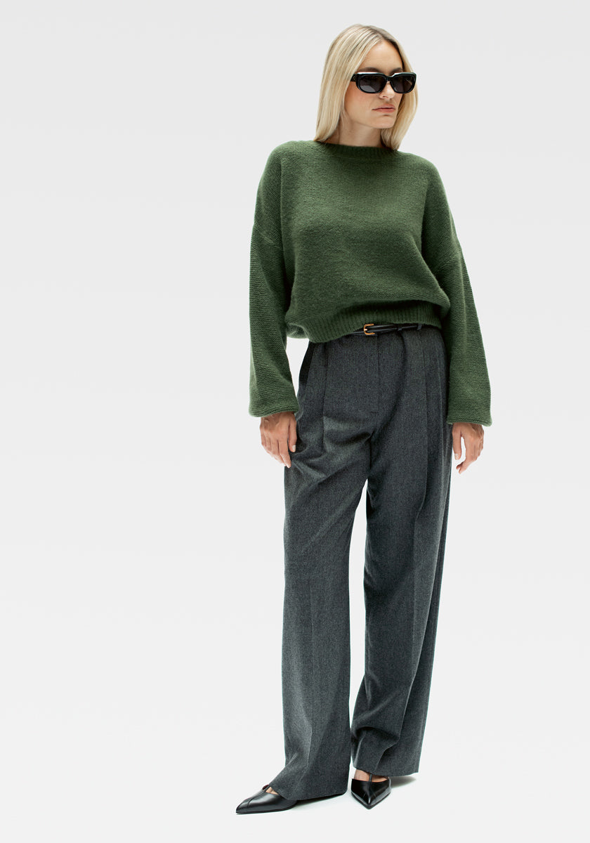 Eine Frau mit glattem, hellblondem Haar und einer entspannten, selbstbewussten Körperhaltung trägt einen Princess Goes Hollywood Kaschmir Strickpullover mit Rundhalsausschnitt und ballonförmigen Ärmeln, eine graue Hose mit hoher Taille und weitem Bein, schwarze spitze Absätze und eine große schwarze Sonnenbrille.