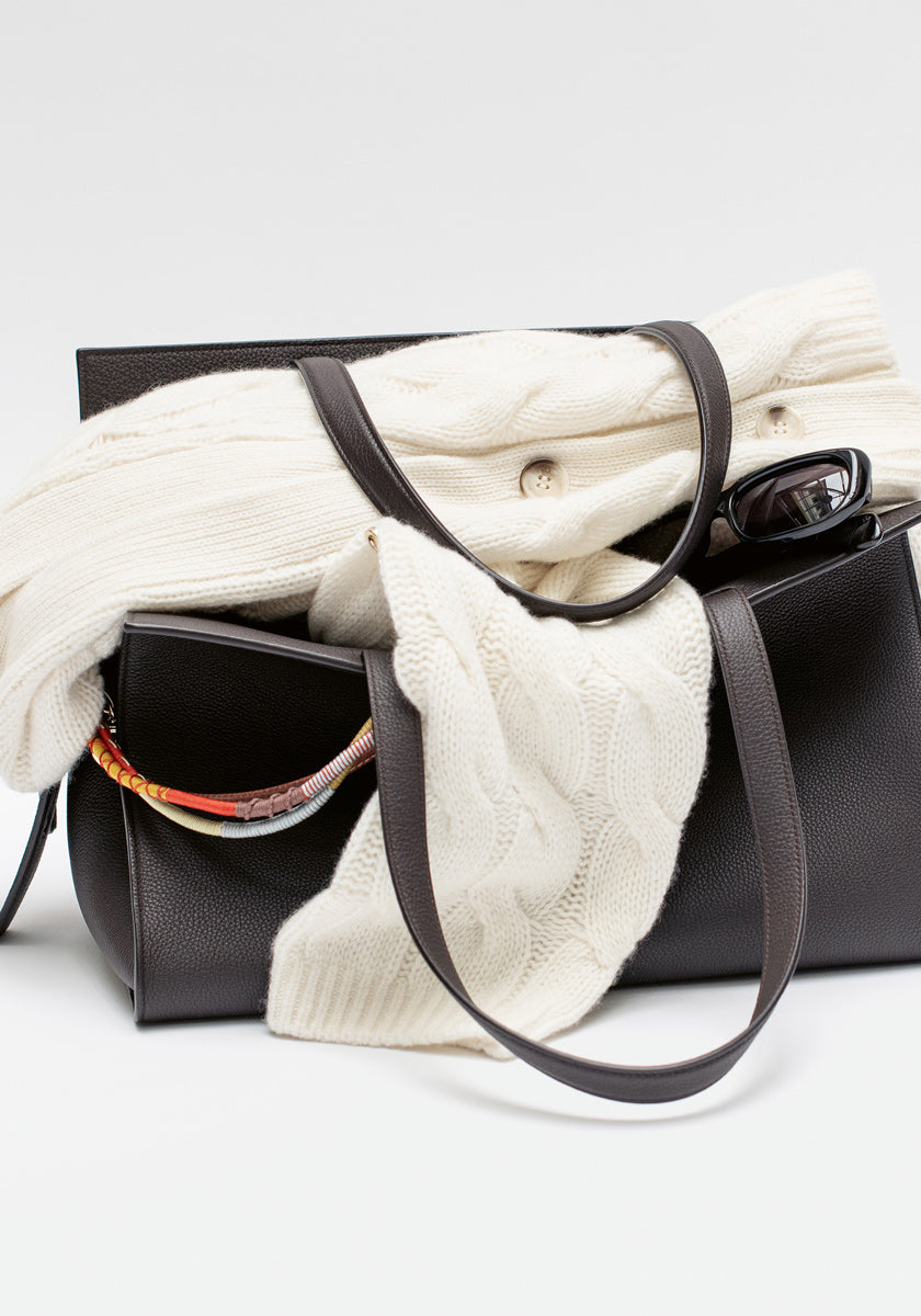 Eine schwarze Lederhandtasche mit langen Henkeln hält eine gefaltete weiße Princess Goes Hollywood Kaschmir Strickjacke in Zopfmuster, eine schwarze Sonnenbrille und einen bunt gemusterten Schal, der über den Rand drapiert ist, vor einem schlichten weißen Hintergrund.