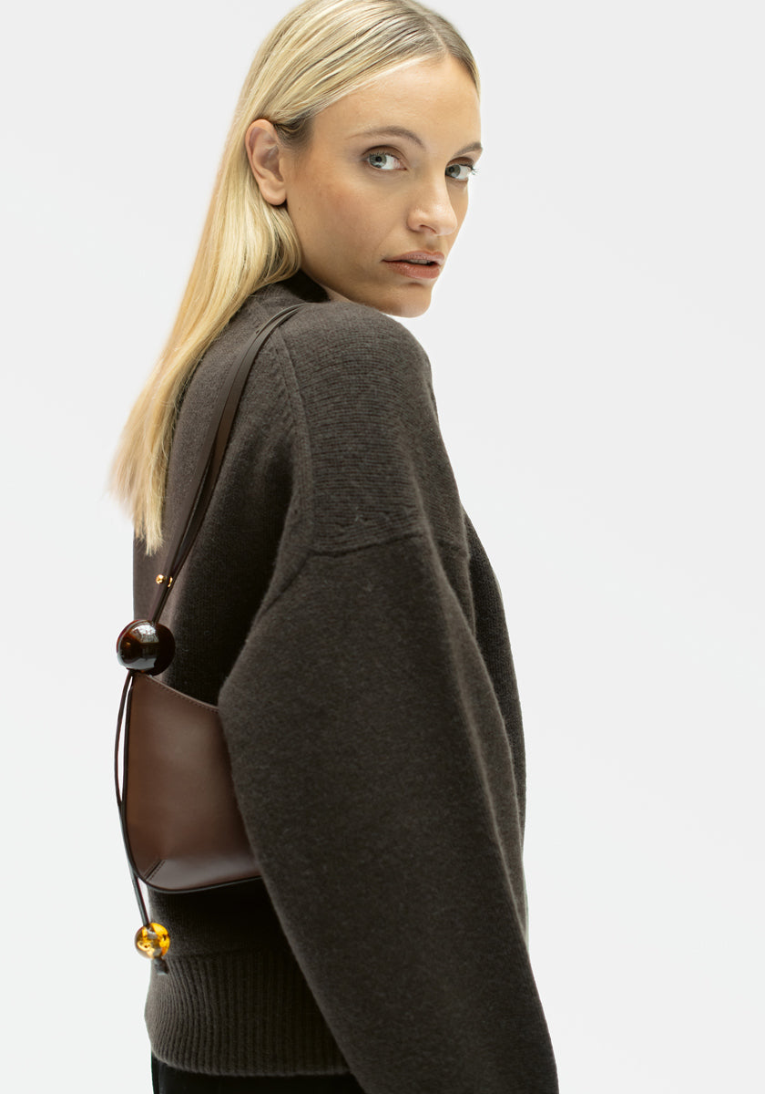 Eine Frau mit glattem blondem Haar modelliert eine lockere, dunkelbraune Kaschmir-Strickjacke von Princess Goes Hollywood und trägt eine kleine braune Umhängetasche mit großen Perlen, während sie vor einem schlichten hellen Hintergrund über ihre Schulter in die Kamera blickt.