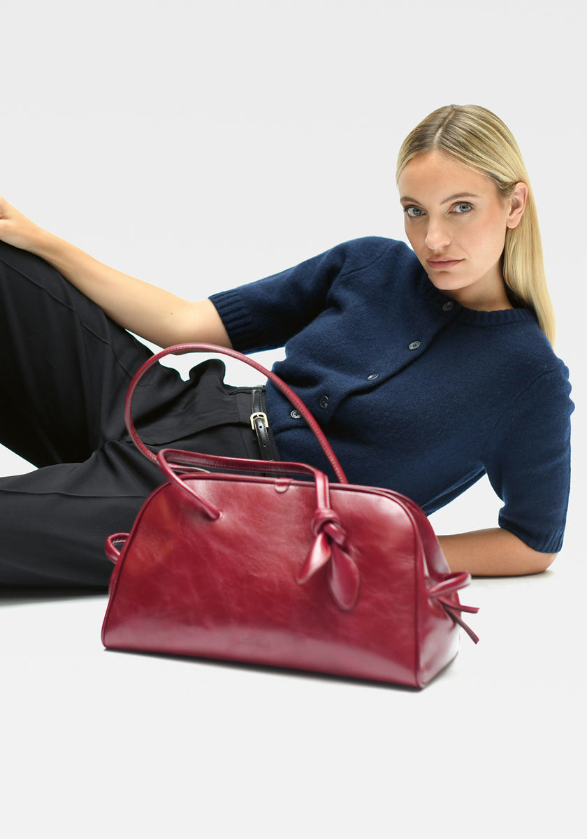 Eine Frau mit langen blonden Haaren trägt einen marineblauen Princess Goes Hollywood Kaschmir Strickpullover mit kurzen Ärmeln und einer schwarzen Hose, die auf einer weißen Fläche ruht. Vorne sticht eine glänzende dunkelrote Lederhandtasche mit geknotetem Seitenteil hervor.