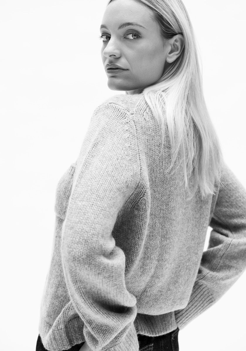 Schwarz-Weiß-Foto einer Frau mit langen blonden Haaren, die über ihre Schulter schaut. Sie trägt eine strukturierte, helle Princess Goes Hollywood Kaschmir Strickjacke mit langen Ärmeln und eine dunkle Hose und steht vor einem schlichten Hintergrund.