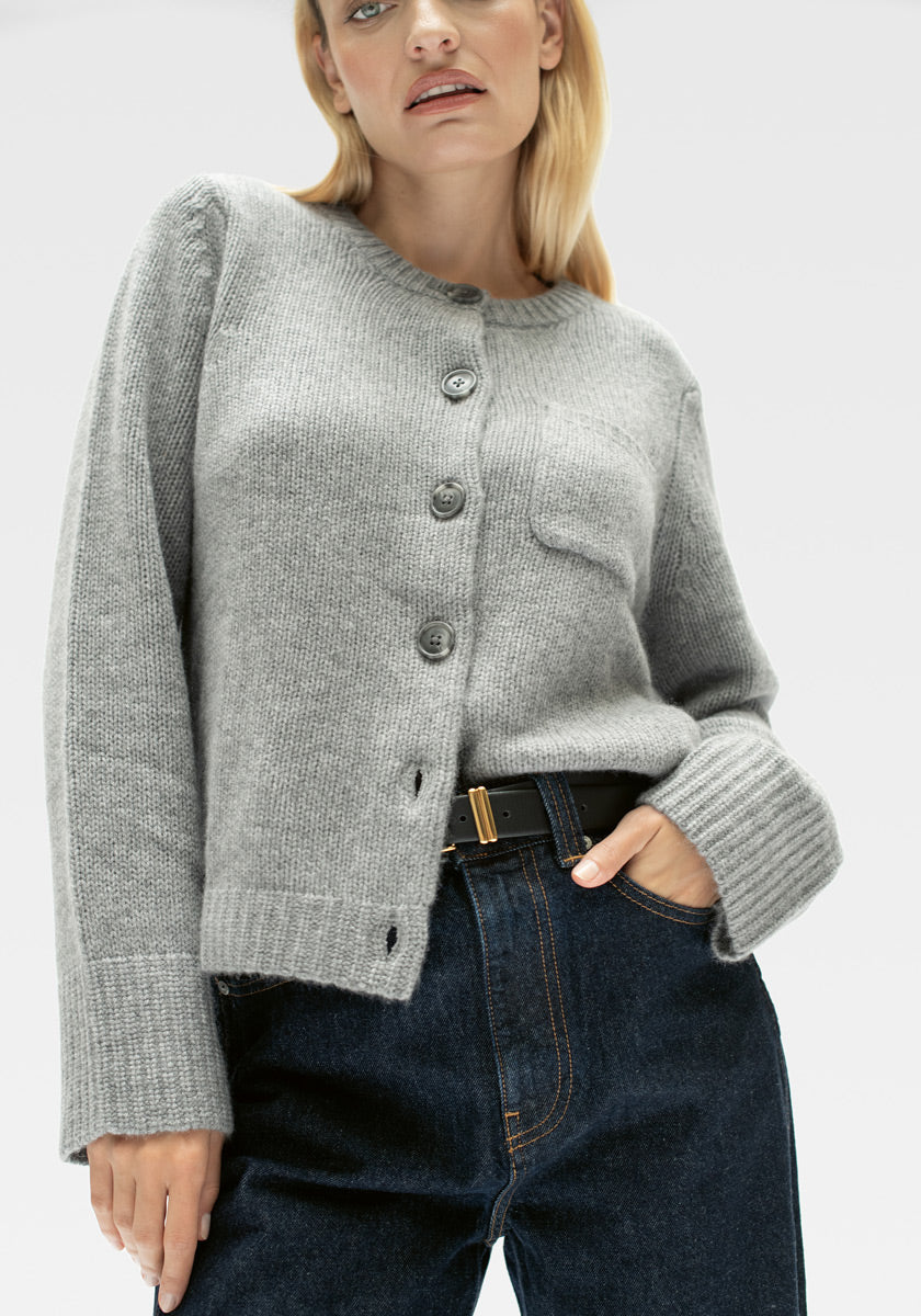 Eine Person trägt eine hellgraue Princess Goes Hollywood Kaschmir Strickjacke mit geknöpfter Vorderseite, Rundhalsausschnitt, Brusttasche und langen Ärmeln, gestylt mit hochtaillierten dunkelblauen Jeans und einem schwarzen Gürtel mit Goldschnalle. Blondes Haar, neutraler Ausdruck, hellgrauer Hintergrund.