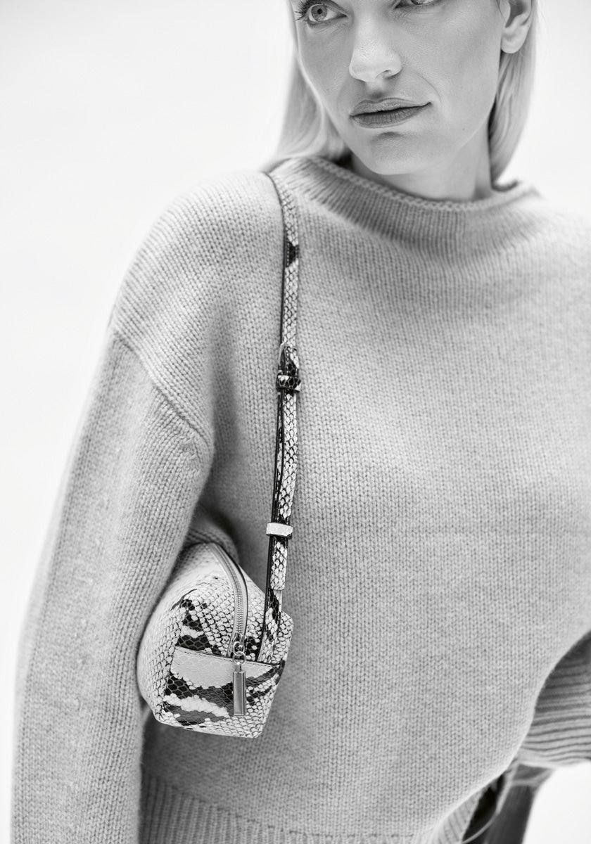 Schwarz-Weiß-Foto einer Frau in einem Princess Goes Hollywood Kaschmir Strickpullover mit Stehkragen und langen Ärmeln. Sie hat glattes, helles Haar und trägt eine kleine, mit Schlangenhaut gemusterte Umhängetasche mit Reißverschluss und Schnalle.