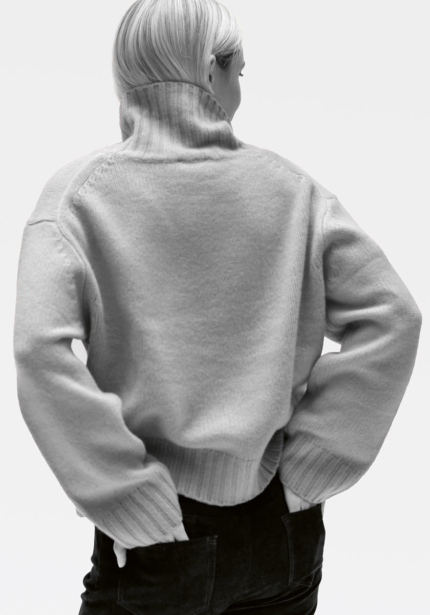 Eine Person mit hellblondem Haar in einem geraden Bob ist von hinten zu sehen und trägt einen klobigen Princess Goes Hollywood Kaschmir Rollkragenpullover mit weiten Ärmeln und eine schwarze Hose. Ihre Hände ruhen auf ihren Hüften vor einem schlichten, hellgrauen Hintergrund.