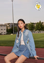 A young woman with straight black hair sits on a railing at an outdoor track, wearing a Princess Goes Hollywood Snoopy & Woodstock-print denim jacket and skirt over a white top. Modern buildings, trees, and the Peanuts 75th Anniversary logo are in the background.