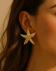 A close-up of a woman’s ear wearing a large, shiny Princess Goes Hollywood Seestern-Ohrstecker with Gelbgold Vergoldung. Her brown hair drapes beside her face, contrasting with her warm skin and the softly lit, neutral background.