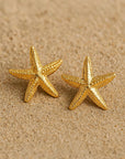 A close-up of two Princess Goes Hollywood Seestern-Ohrstecker aus Edelstahl mit Gelbgold Vergoldung rests on sand, evoking a beach vibe. Placed side by side, they showcase their shiny finish and detailed design.