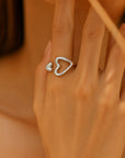 A close-up of a hand wearing a Princess Goes Hollywood Edelstahlring Damen in Herzform: a silver open ring featuring two heart shapes, ideal as a Geschenk mit Herz, set against a softly blurred beige background.
