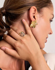 A woman with brown hair in a ponytail touches her ear, displaying a gold flower-shaped earring and two Princess Goes Hollywood Ring Edelstahl (one gold, one silver) with Herz Design. Perfect as a gift for women.
