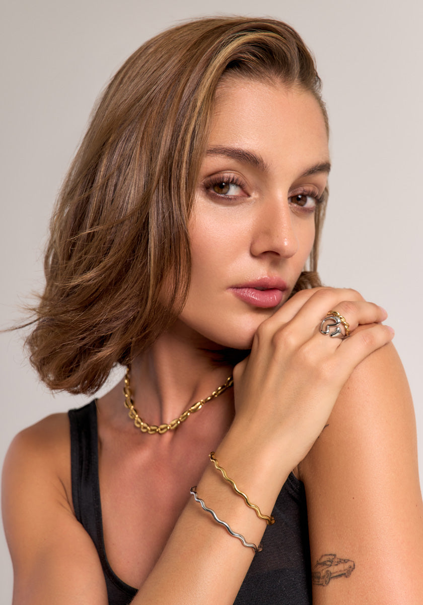 A woman with short, wavy brown hair poses against a neutral background. She wears Princess Goes Hollywood gold and silver jewelry, including a filigranes Schmuckstück und verstellbarer Ring in Edelstahl Gelbgold, with natural makeup and a black tank top.