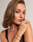 A woman with short, wavy brown hair poses against a neutral background. She wears Princess Goes Hollywood gold and silver jewelry, including a filigranes Schmuckstück und verstellbarer Ring in Edelstahl Gelbgold, with natural makeup and a black tank top.