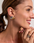 A light-skinned woman with brown hair in a ponytail smiles, wearing sculptural gold and silver earrings and a matching Ring Edelstahl by Princess Goes Hollywood—an ideal Geschenk für Frauen. The background is plain and light-colored.