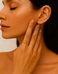 A woman with smooth, tan skin and long dark hair gently touches her face, displaying a Princess Goes Hollywood adjustable stainless steel ring with yellow gold plating. Her closed eyes and natural makeup highlight her serene, elegant features.