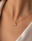 Eine Nahaufnahme einer Person, die eine zarte Goldkette von Princess Goes Hollywood trägt, mit einem Anhänger in Form des Buchstabens „M“, der mit funkelnden Steinen besetzt ist. Dazu trägt sie ein helles Oberteil mit offenem Kragen; der Hintergrund ist sanft unscharf.