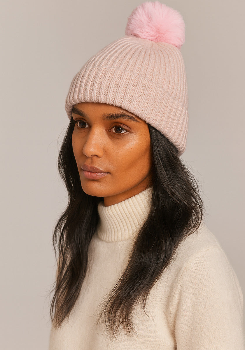 A woman with long dark hair wears a cream turtleneck and a pale pink ribbed cashmere beanie with a fluffy pom-pom by Princess Goes Hollywood, looking slightly to the side against a neutral beige background.
