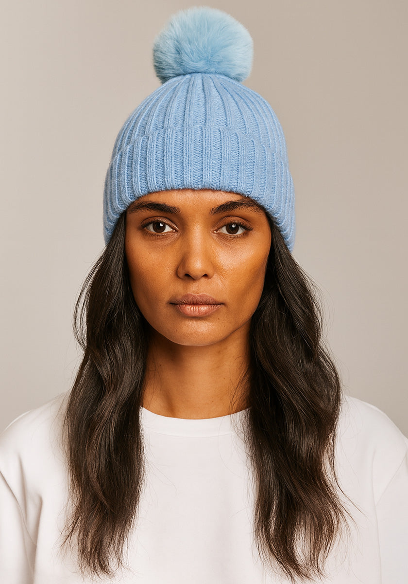 A woman with long, wavy dark hair wears a light blue Princess Goes Hollywood Bommelmütze featuring a fluffy pom-pom. She faces forward in a plain white shirt against a simple light beige background, her expression neutral.