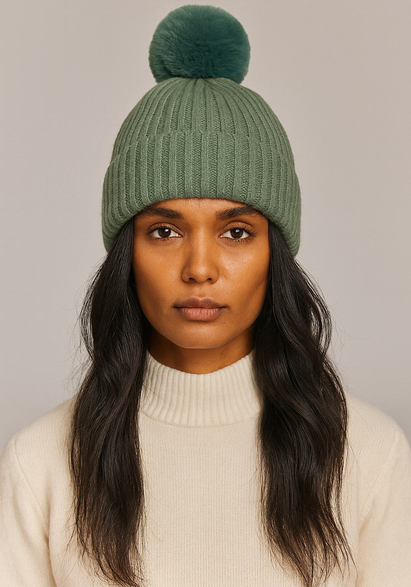 A woman with long, wavy dark hair wears a sage green Princess Goes Hollywood Rippstrick Kaschmir Mütze with a large pom-pom. She has a neutral expression and is dressed in a cream ribbed turtleneck sweater against a light gray background.