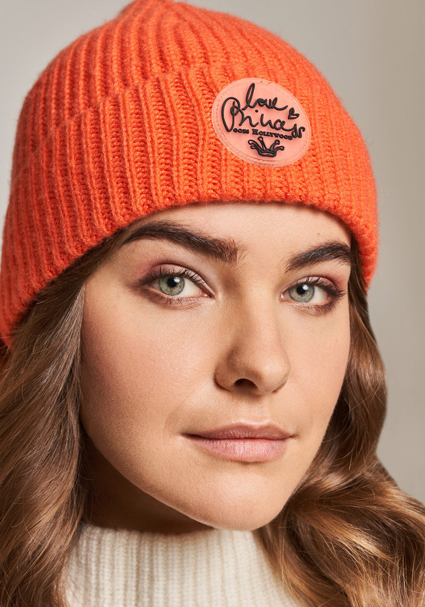 A person with wavy brown hair and light eyes wears an orange knitted beanie with a Love Brings Some Lawood badge and a white ribbed sweater by Princess Goes Hollywood. The background is neutral.
