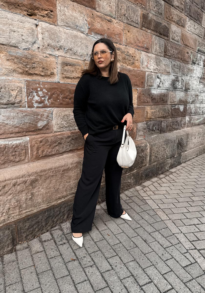 A woman wearing a Princess Goes Hollywood black sweater with round neckline, black wide-leg pants, and white pointed-toe shoes stands by a stone wall. She carries a white handbag, wears glasses, and gazes confidently to the side.