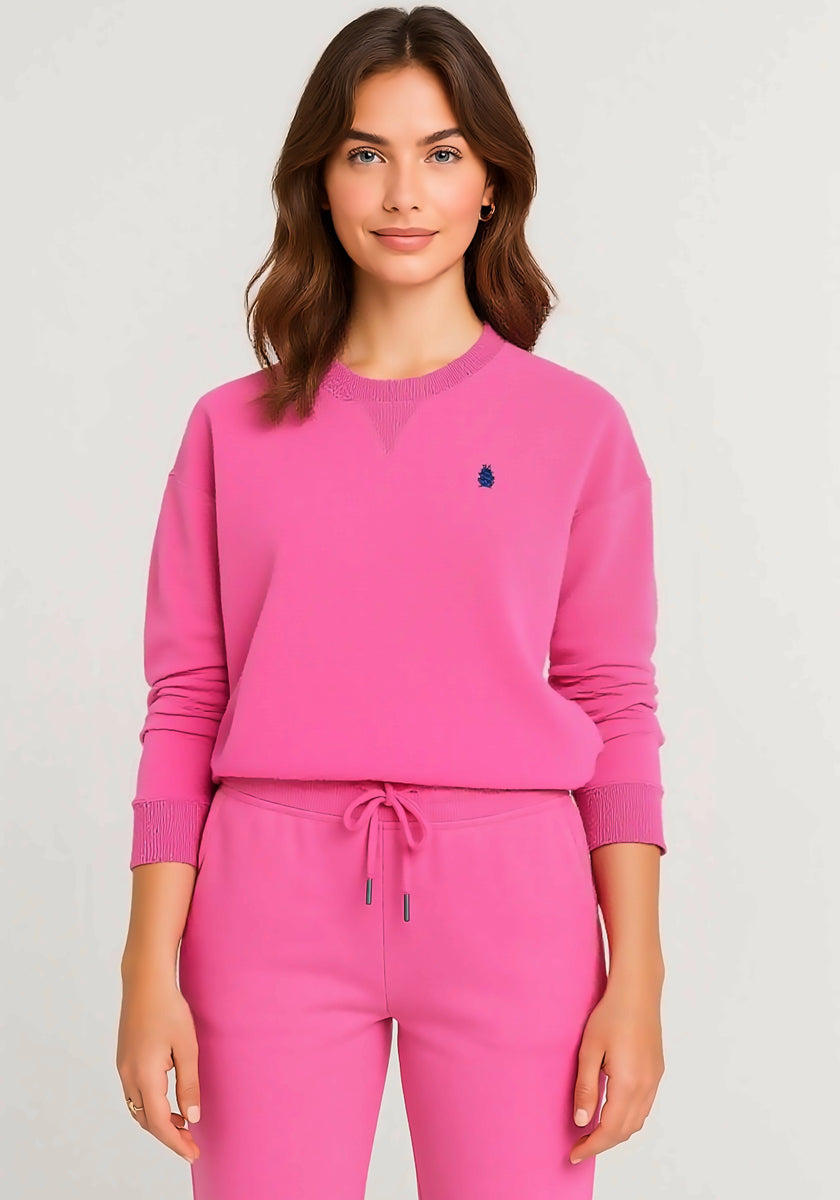 A woman with wavy brown hair wears a bright pink Princess Goes Hollywood fleece sweatpullover with a small dark blue embroidered logo on the left chest and matching drawstring sweatpants, standing against a plain light background.