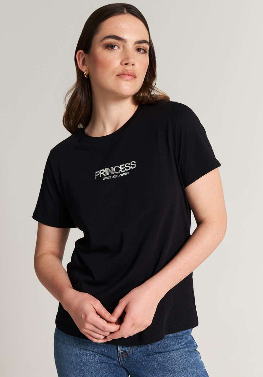 A woman with long brown hair wears a black cotton Princess Goes Hollywood T-shirt with a crew neck and blue jeans. She stands against a plain light background, looking forward neutrally, hands gently clasped at her waist.