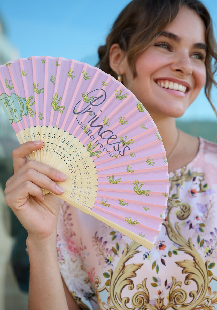 A smiling woman with wavy brown hair holds the Handfächer Princess by Princess Goes Hollywood, featuring pink and purple tones with green frogs and Princess text. She pairs it with a pastel floral blouse for a fashionable outdoor look.