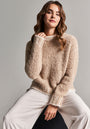 A woman with wavy brown hair sits confidently on a stool against a neutral background, wearing the Beiger Grobstrickpullover mit Kontrastnähten from Frogbox by Princess and loose-fitting white pants, looking directly at the camera.