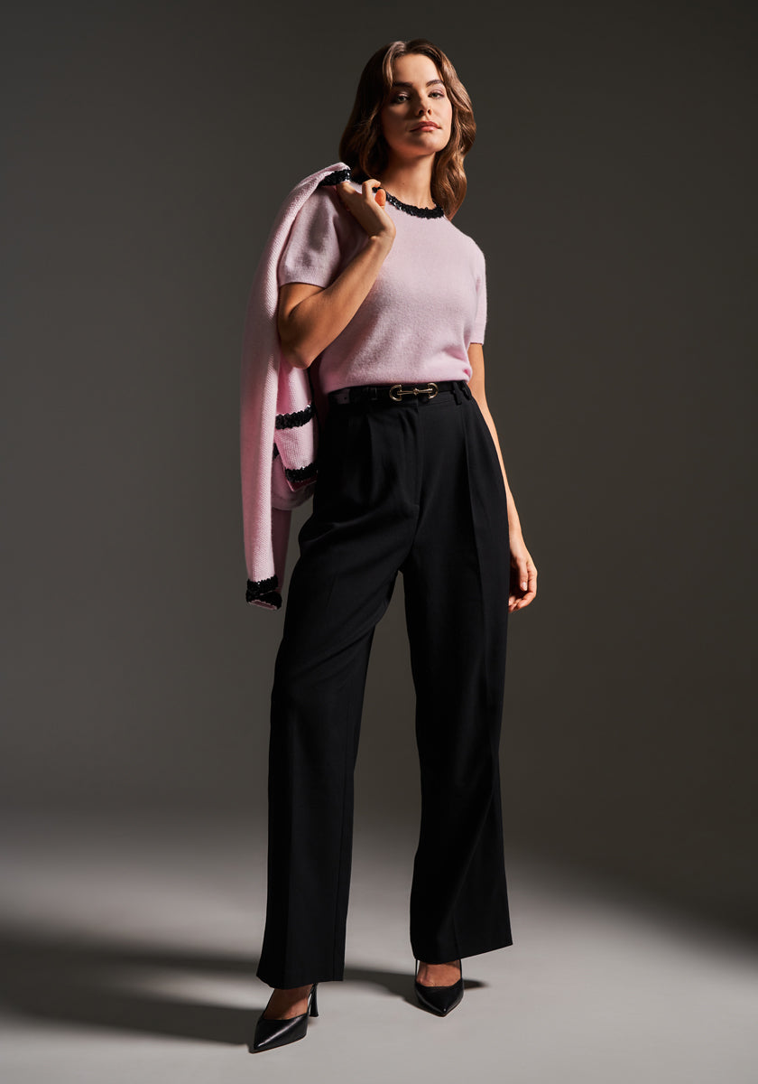 A woman poses confidently against a dark backdrop in a short-sleeved pink Princess Goes Hollywood Wolle sweater with dark trim, black high-waisted trousers, pointed heels, and a matching pink Princess Goes Hollywood Kaschmir sweater draped over one shoulder.