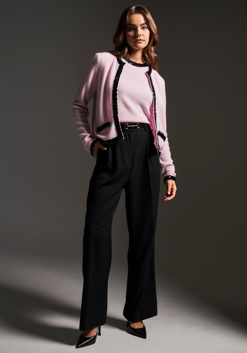 A woman stands confidently, wearing a light pink cardigan with black trim over a matching top, paired with high-waisted black trousers and black pointed-toe heels. The background is plain and dark, drawing focus to her stylish, modern outfit.