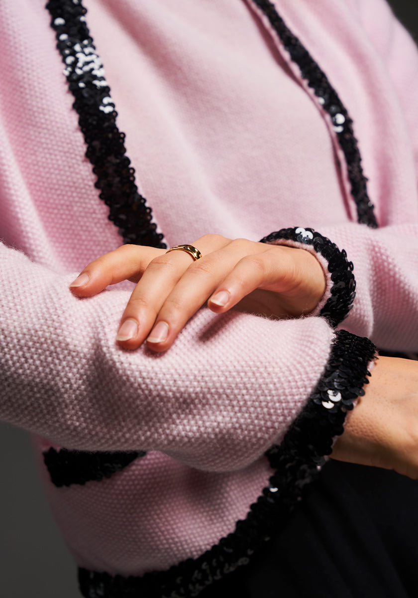 A close-up of hands resting on a pink Princess Goes Hollywood Kaschmir sweater with black sequin detailing. The person wears a gold ring and has neatly manicured, natural nails. The background is dark and softly blurred.