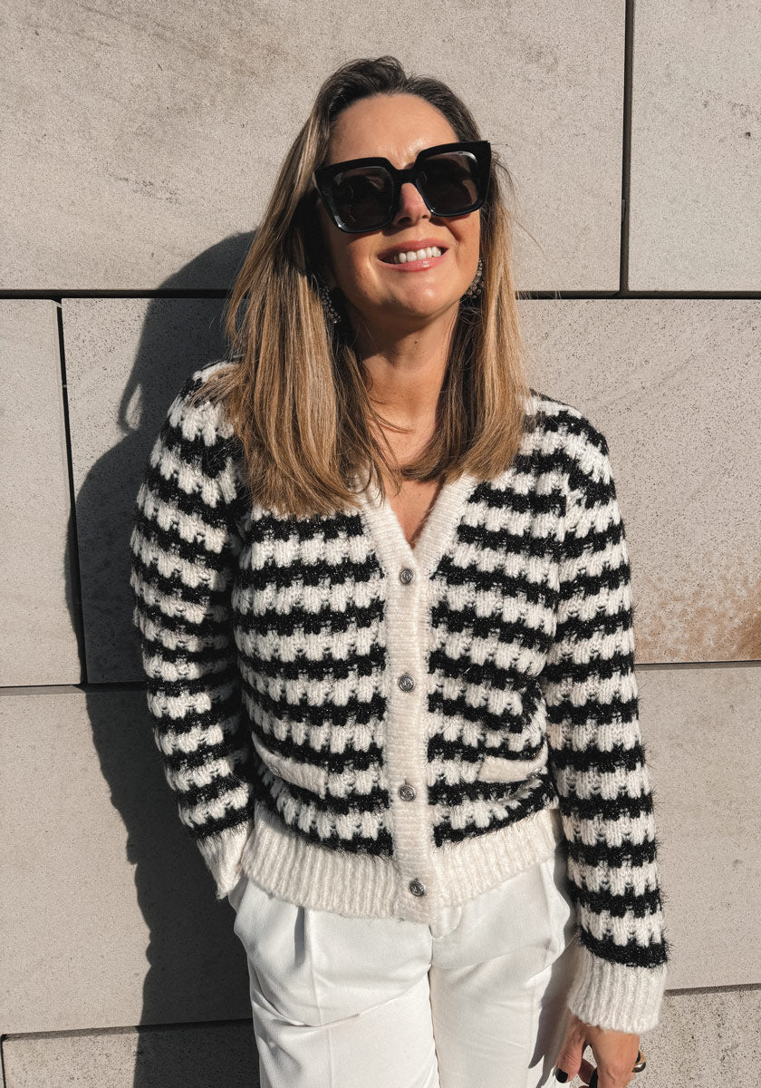 A woman with straight, shoulder-length hair wears large black sunglasses and a black-and-white Princess Goes Hollywood Grobstrickjacke with silver buttons, plus white pants. She smiles in front of a beige stone wall under bright sunlight.
