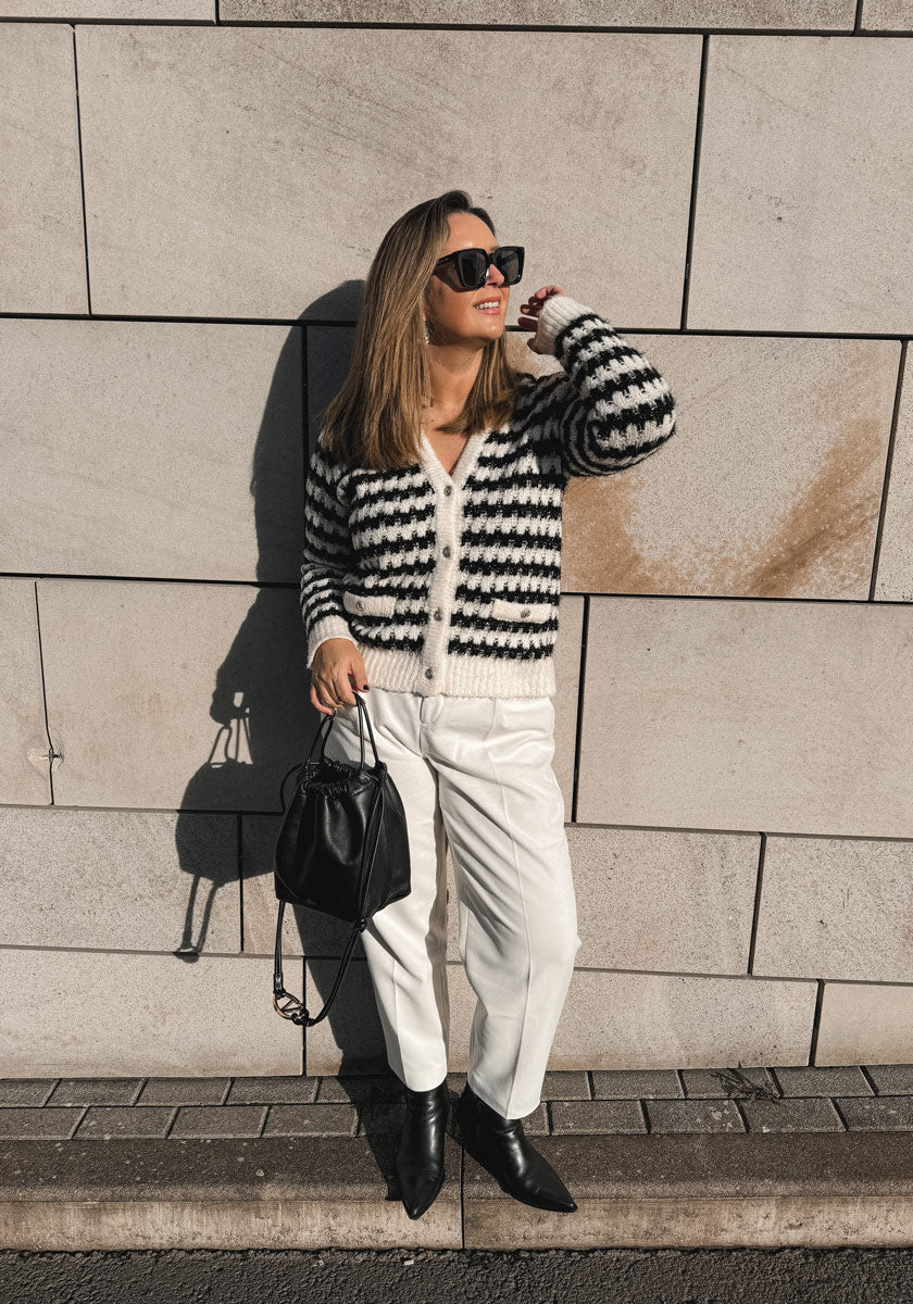 A woman smiles in sunlight against a tiled wall, wearing sunglasses, white pants, black pointed boots, and a black-and-white Grobstrickjacke with Lurexfäden by Princess Goes Hollywood. She holds a black handbag as warm tones highlight the scene.