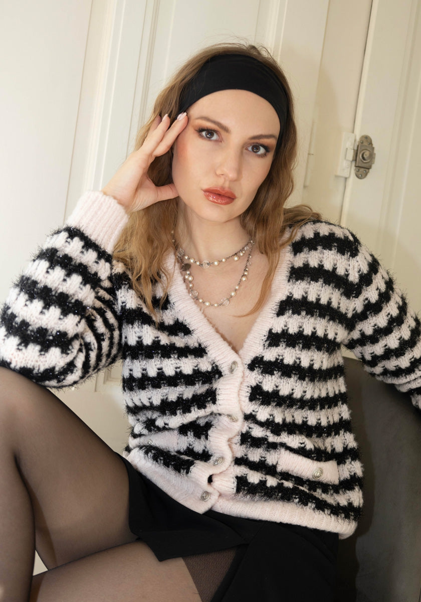 A woman with wavy light brown hair wears a black headband, pearl necklace, Princess Goes Hollywood black-and-white striped Grobstrickjacke, black skirt, and sheer tights. She sits by a light-colored door, resting her head on one hand and gazing at the camera.