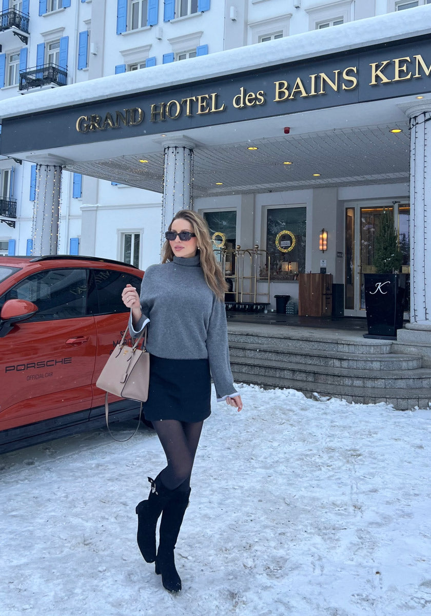 A woman in sunglasses and a gray Princess Goes Hollywood Rollkragen sweater with subtle sparkles, paired with a black mini skirt and boots, stands on snowy ground near a red Porsche by the GRAND HOTEL des BAINS KEMPINSKI.