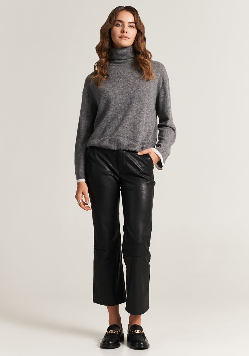 A woman with loose wavy brown hair stands confidently against a light background, wearing a gray Princess Goes Hollywood Rollkragen sweater, black leather pants, and black loafers, one hand in her pocket.