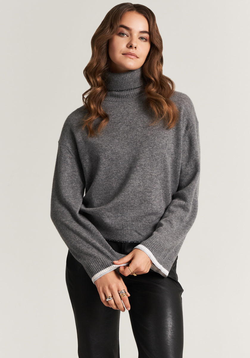 A woman with wavy brown hair wears a Princess Goes Hollywood graugrauer Rollkragen-Feinstrickpullover in Kaschmir-Mix with light cuffs and black leather pants, standing against a plain background with her hands loosely clasped.