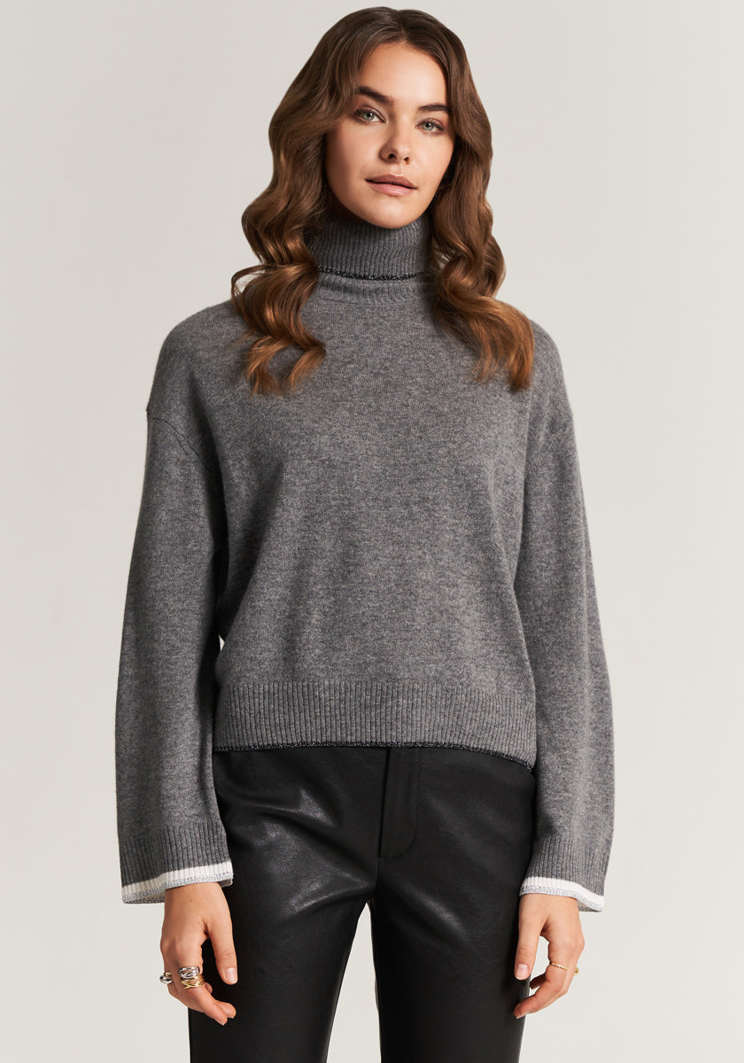 A woman with wavy brown hair wears a gray Princess Goes Hollywood Rollkragen sweater with ribbed cuffs and hem plus subtle Glitzerdeko, standing against a plain light background with arms relaxed and a neutral expression.