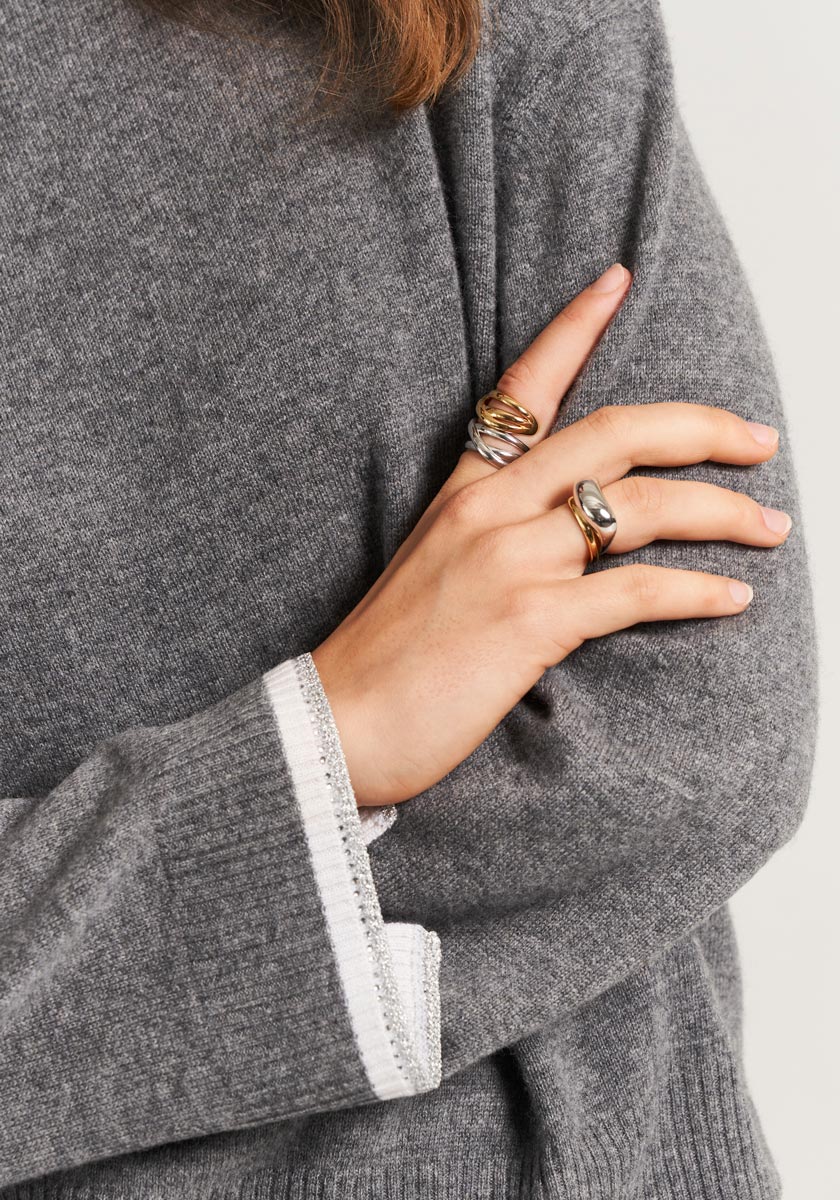 A person wearing a soft grey Princess Goes Hollywood knit Rollkragen sweater with white sleeve trim crosses their arms, showing two chunky metallic rings—one gold, one silver. Only their hand, arm, and part of their torso are visible.