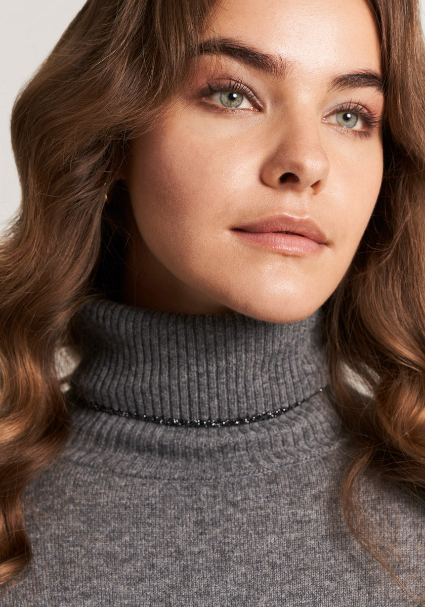 A young woman with wavy light brown hair wears a Princess Goes Hollywood Feinstrickpullover aus Kaschmir Mischung mit Rollkragen. She has clear skin, light eyes, and natural makeup, gazing calmly to the side against a softly lit background.