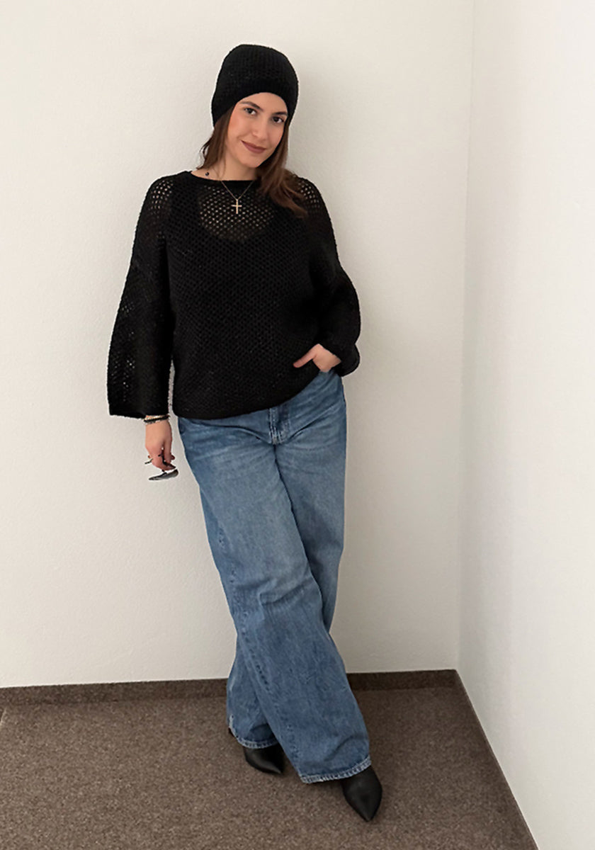 A woman stands in a corner, smiling slightly, dressed in a black Princess Goes Hollywood Merino knit sweater, matching Strickmütze, blue wide-leg jeans, and black pointed shoes. One hand is in her pocket and she holds eyeglasses against white walls.