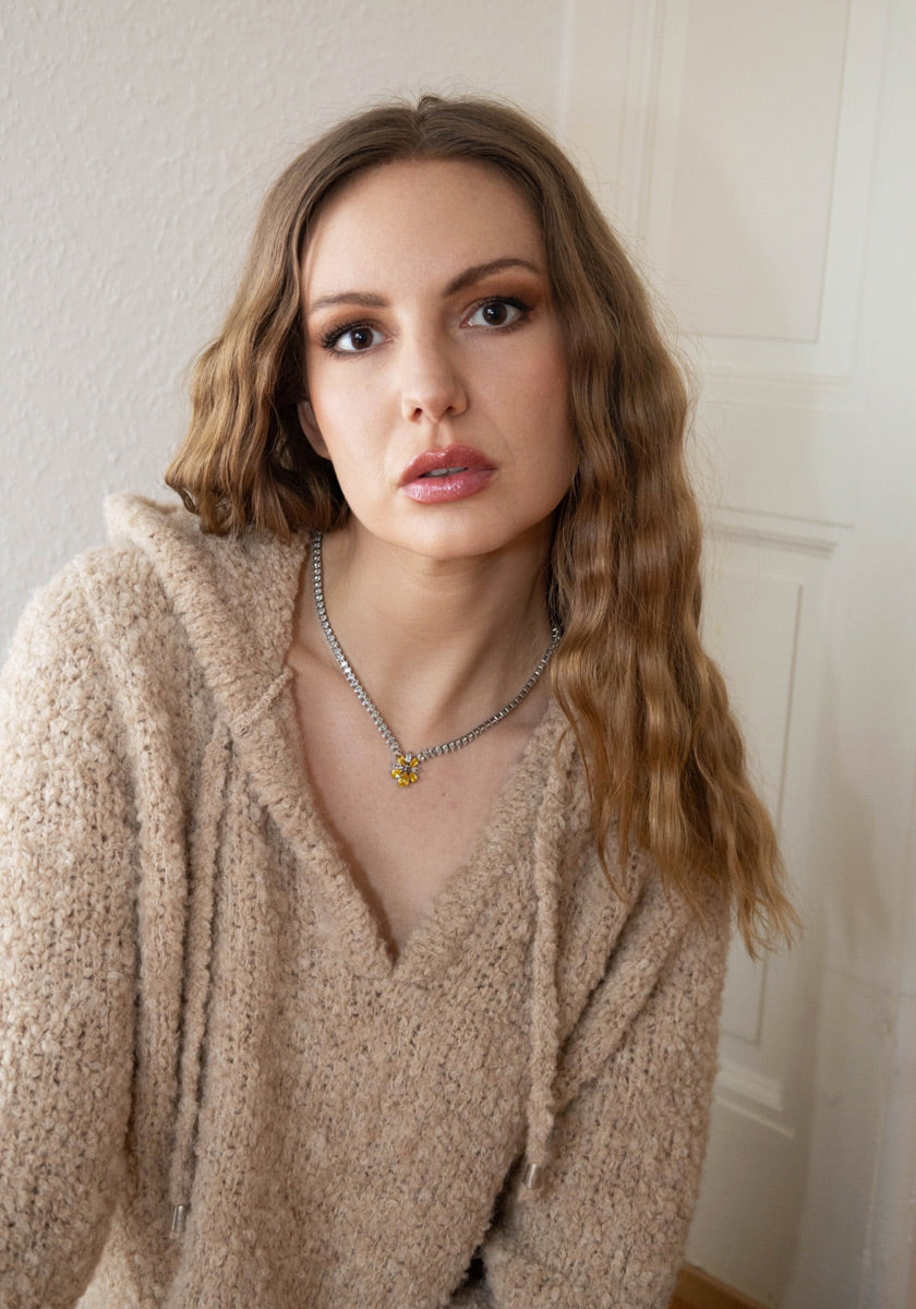A young woman with long, wavy brown hair wears a beige Grobstrick-Hoodie with Kontrastnähte from Frogbox by Princess and a chunky silver necklace with a yellow pendant. She faces the camera, seated before a cream-colored wall and door.