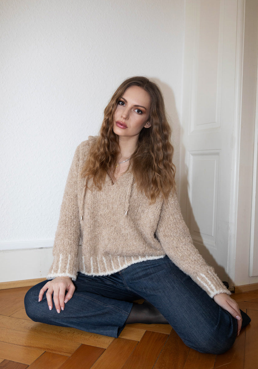 A young woman with wavy light brown hair sits on a wooden floor against a white wall and door, wearing a beige Grobstrick-Hoodie with white contrast stitching by Frogbox by Princess and blue jeans. She looks directly at the camera, one hand on the floor.