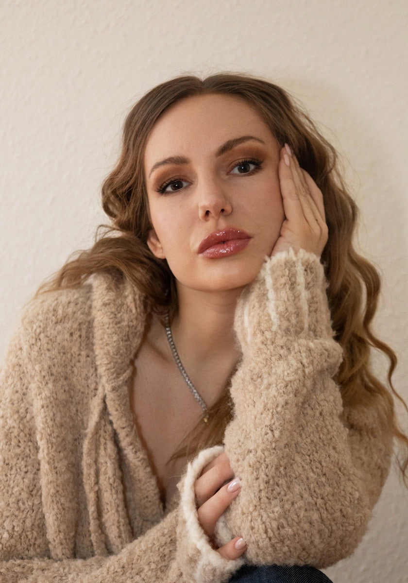 A woman with wavy light brown hair rests her face on her hand, gazing at the camera. Shes wearing a cozy beige Froxbox by Princess Alpaka Grobstrick-Hoodie mit Kontrastnähten. Subtle makeup and an off-white wall create a calm, intimate mood.