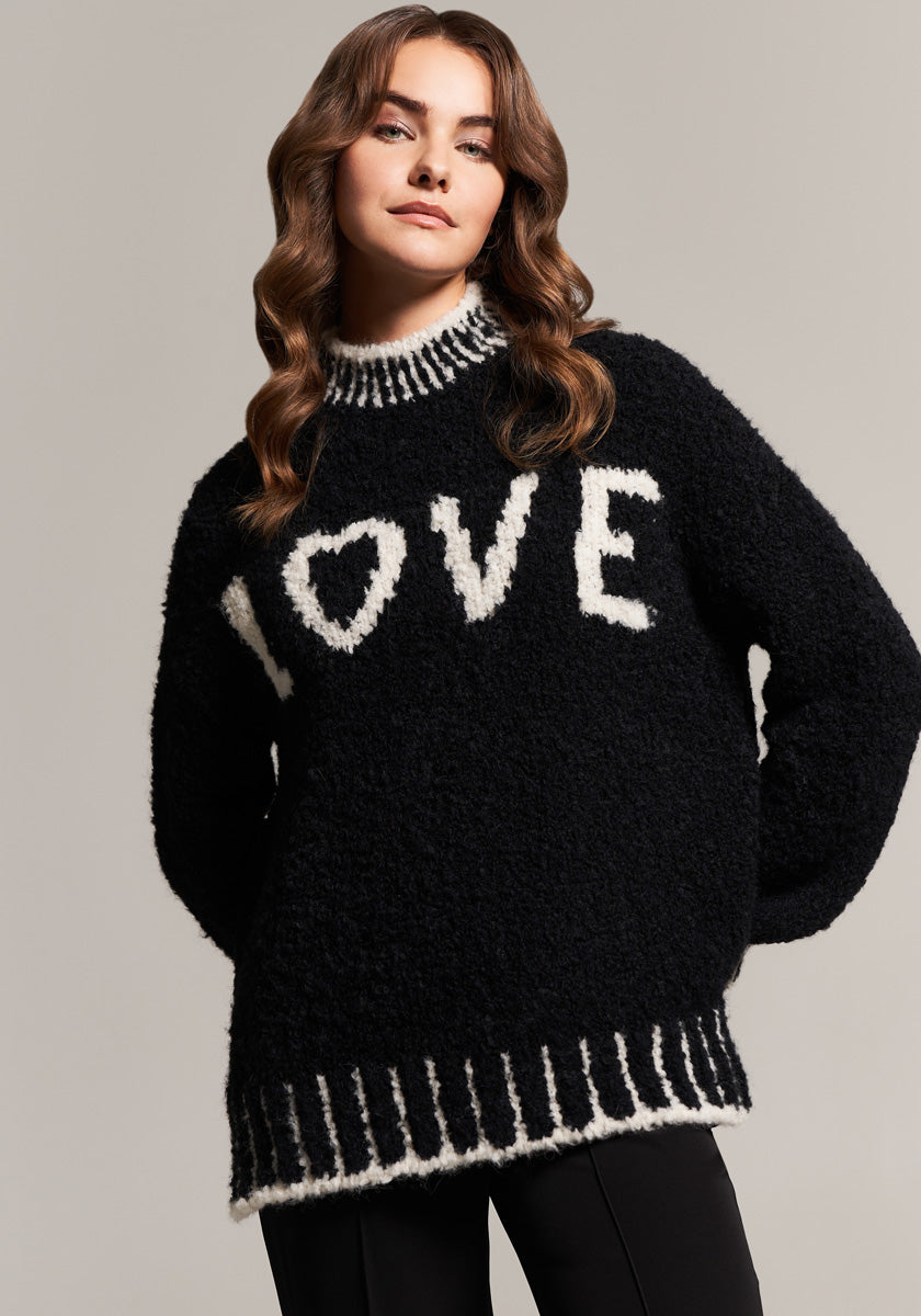 A woman with wavy brown hair wears a Frogbox by Princess Grobstrickpullover aus Alpaka Wolle Mischung in fuzzy black with white contrast trim and bold “LOVE” lettering, standing confidently against a plain gray background.